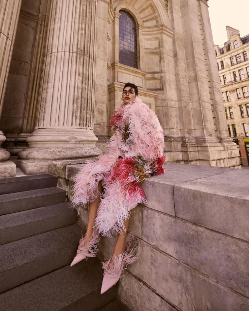 Fashion photography featuring a model in a pink feathered coat and shoes, posing on steps in front of a stone building with pillars.