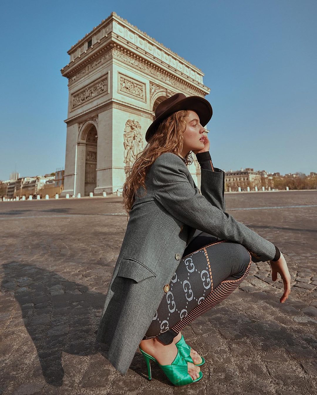 Adult female poses in hat, jacket, and printed leggings with the Arc de Triomphe monument in the background on a sunny day.