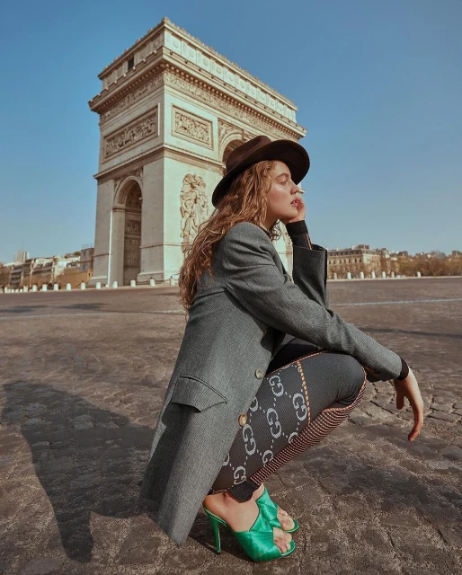 Adult female poses in hat, jacket, and printed leggings with the Arc de Triomphe monument in the background on a sunny day.