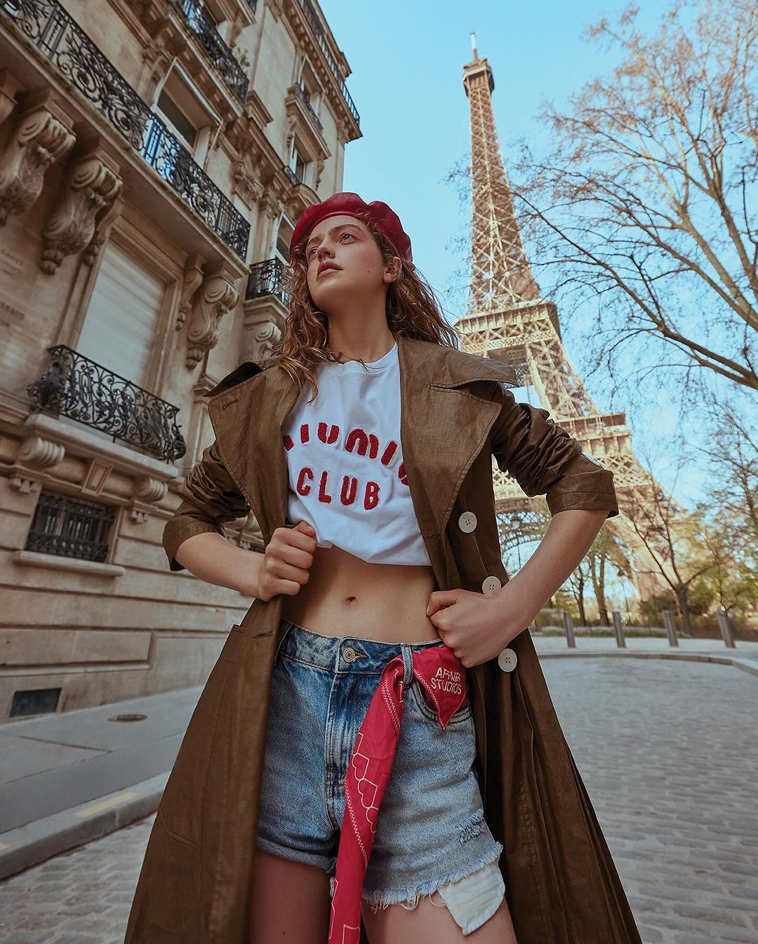 Young woman in trench coat and beret poses in front of the Eiffel Tower, Paris, wearing denim shorts, with architectural details.