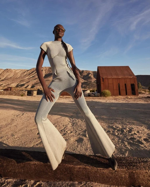 Fashion shot of a Black female model in a grey knit outfit and sunglasses posing outdoors in front of an old rusted building and desert landscape.