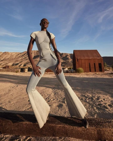 Fashion shot of a Black female model in a grey knit outfit and sunglasses posing outdoors in front of an old rusted building and desert landscape.