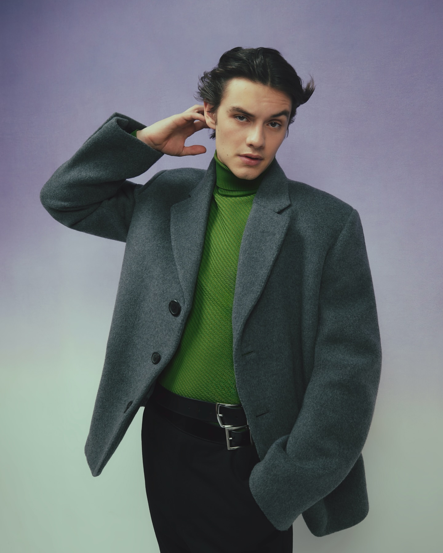 Man in grey coat and green turtleneck poses against a lilac background in a fashion portrait, looking at the camera with hand on head.