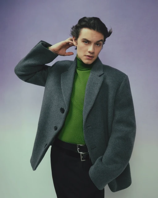 Man in grey coat and green turtleneck poses against a lilac background in a fashion portrait, looking at the camera with hand on head.