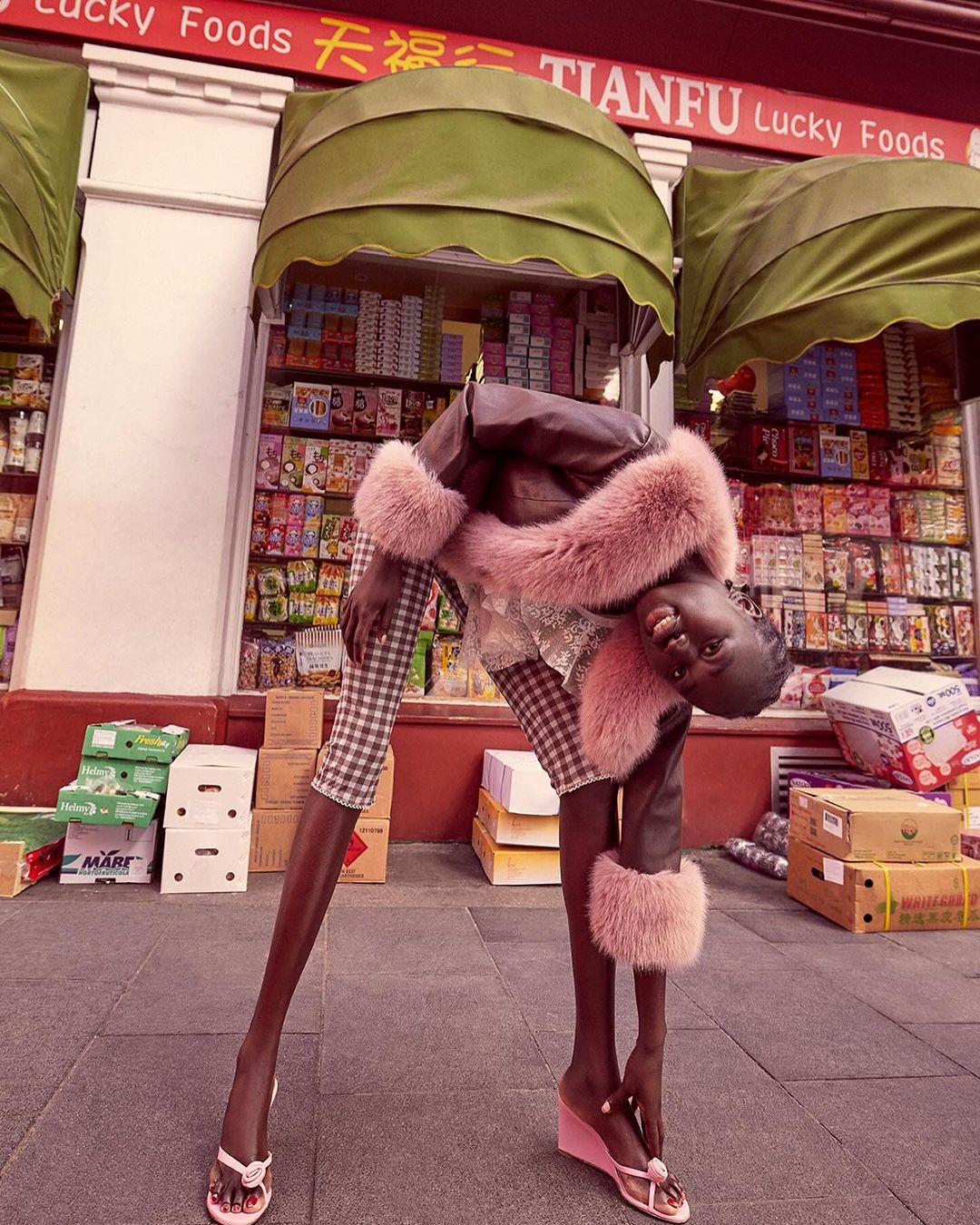 Woman in a brown coat with fur trim and plaid pants poses outside of a Lucky Foods storefront with boxes stacked nearby on the sidewalk.