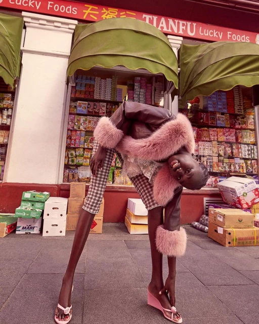 Woman in a brown coat with fur trim and plaid pants poses outside of a Lucky Foods storefront with boxes stacked nearby on the sidewalk.