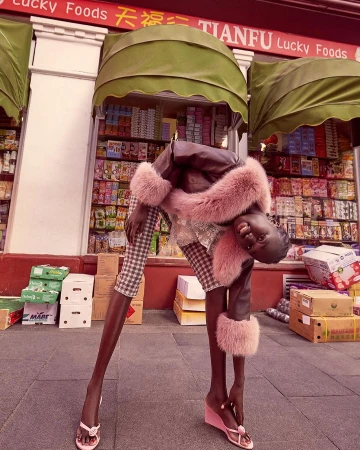 Woman in a brown coat with fur trim and plaid pants poses outside of a Lucky Foods storefront with boxes stacked nearby on the sidewalk.