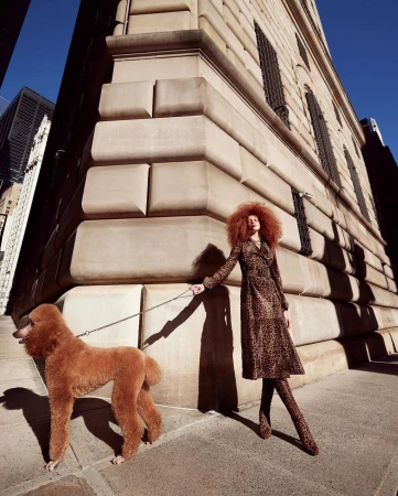 A woman in a patterned coat walks a standard poodle on a city sidewalk, with tall buildings in the background on a sunny day.