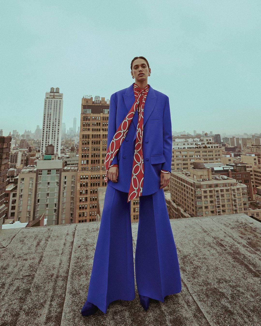 Woman in blue suit and red scarf poses on a rooftop with a cityscape background. Fashion editorial photography with overcast lighting.