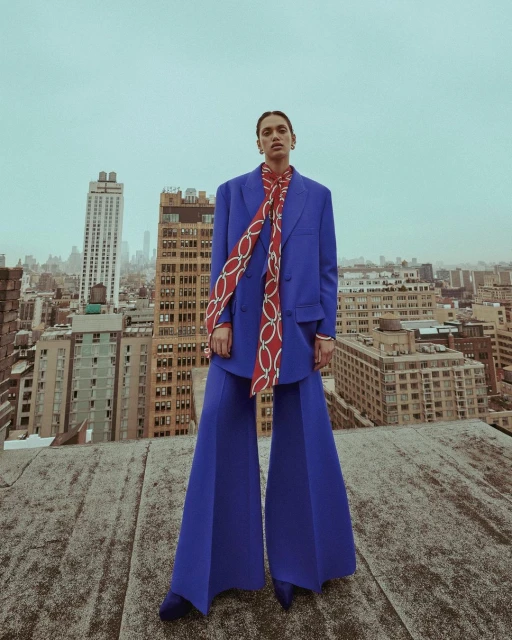 Woman in blue suit and red scarf poses on a rooftop with a cityscape background. Fashion editorial photography with overcast lighting.