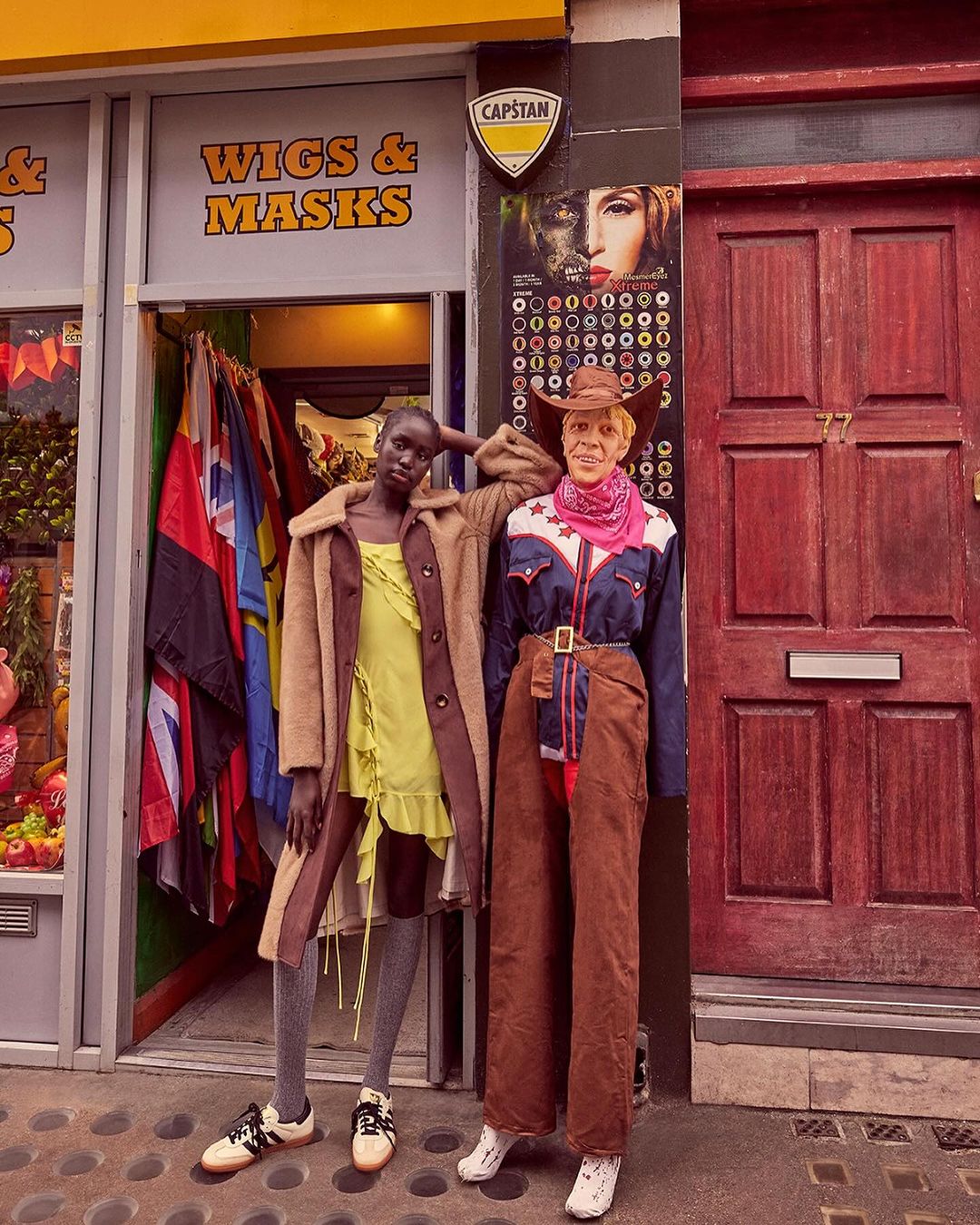 An adult female poses next to a mannequin in front of a Wigs & Masks store. The woman wears a yellow dress and the mannequin is dressed as a cowboy.