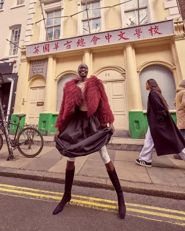 A smiling black model in a textured burgundy faux fur jacket and dark skirt posing outside a London school building by photographer Carla Guler