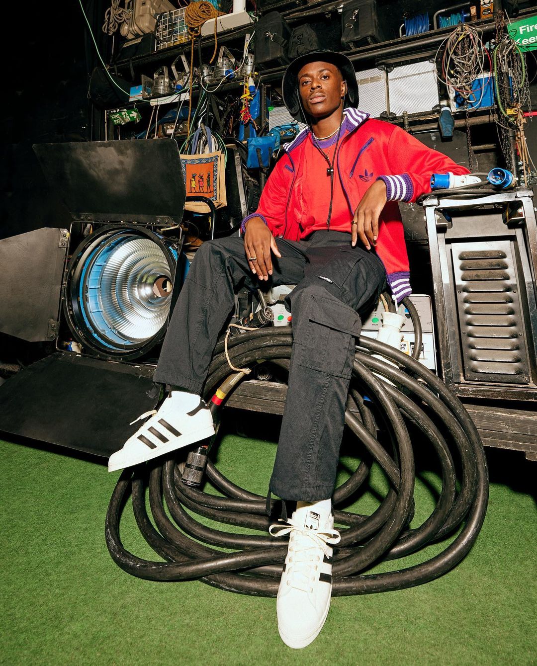 Young man in a red Adidas tracksuit and bucket hat sitting on stage lighting equipment wearing white sneakers with black stripes.