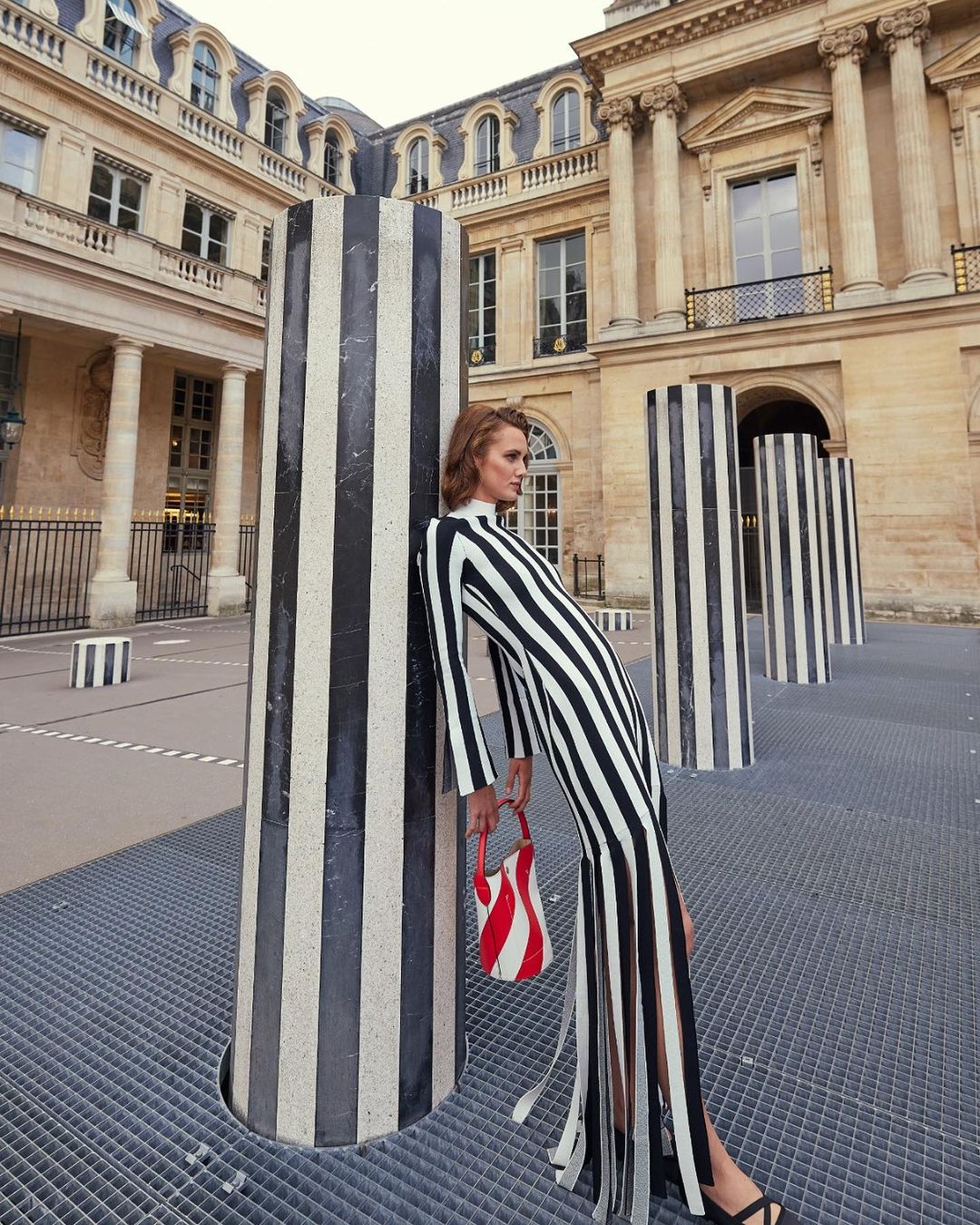 Fashionable woman in striped dress posing by striped columns at the Palais Royal, Paris with historical architecture in the background.