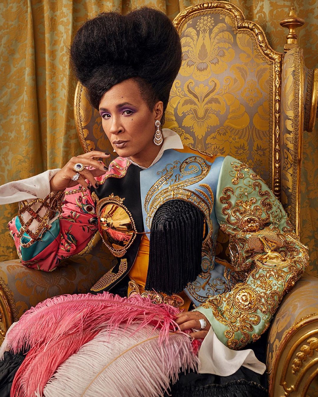 A regal woman with a high afro sits in an ornate gold chair, wearing a colorful jacket with feathers. Rich gold and grey patterned background.