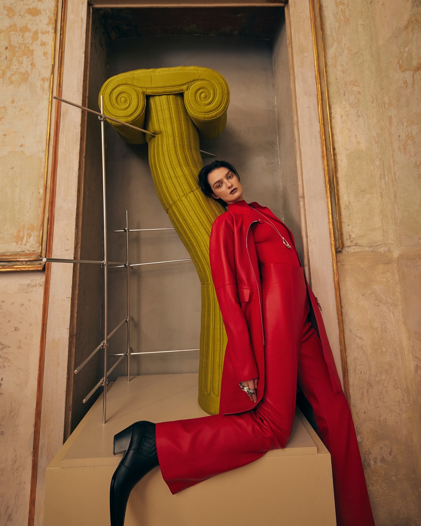 Elegant woman in a red suit poses next to a leaning yellow ionic column inside an ornate room with classical architectural details.