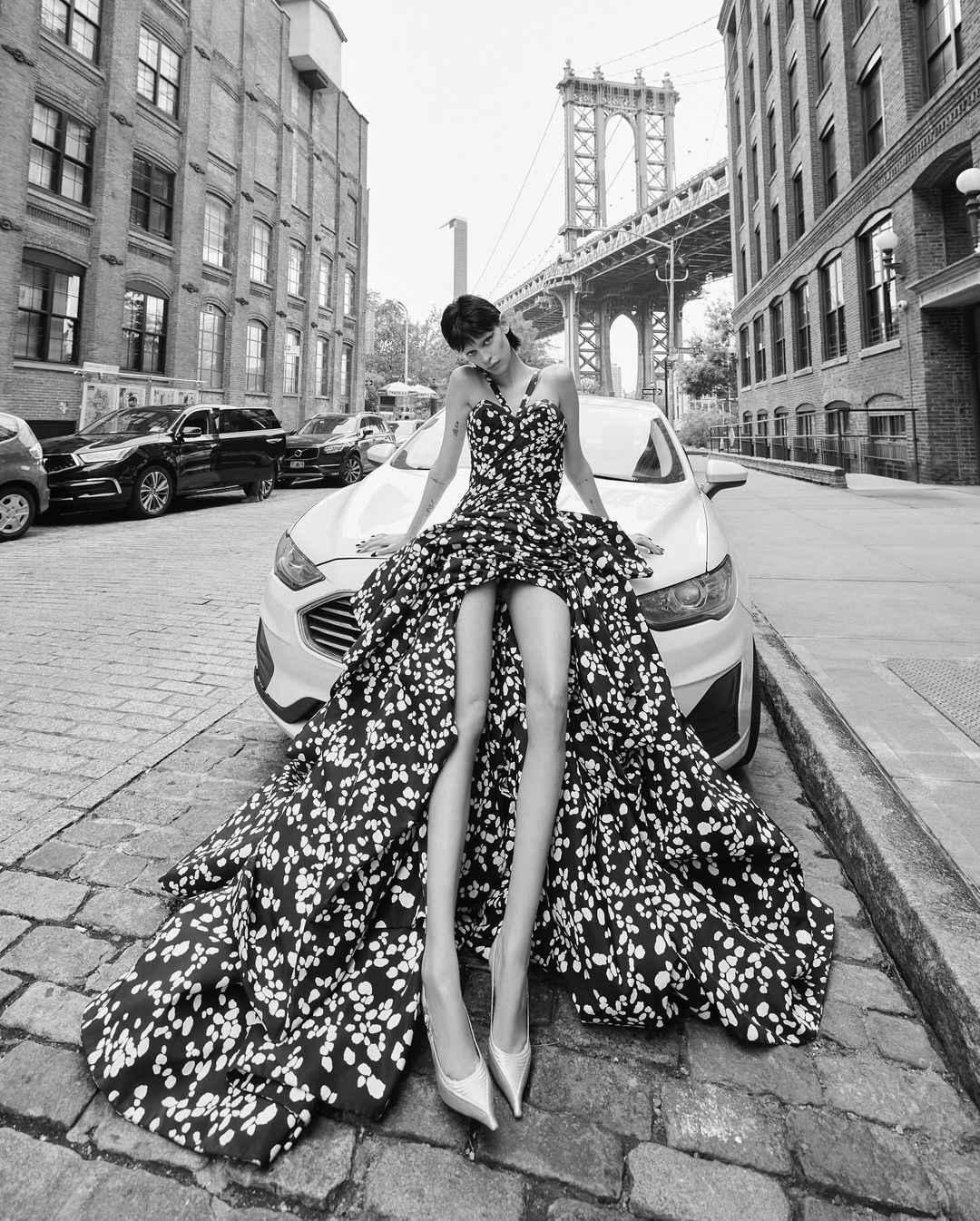 Black and white fashion shot of a woman in a floral dress sitting on a car, urban setting, bridge in background, high fashion mood.