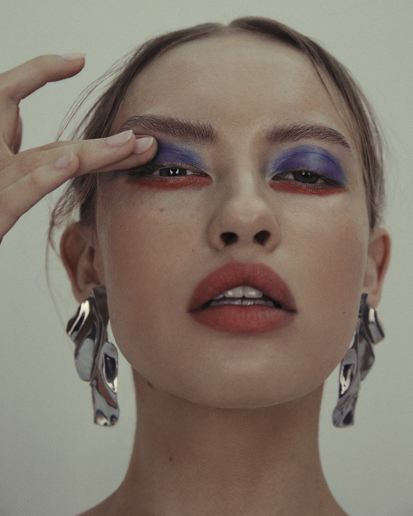 Close-up beauty shot of a woman with red lipstick and dramatic blue and red eye makeup, wearing large silver statement earrings.