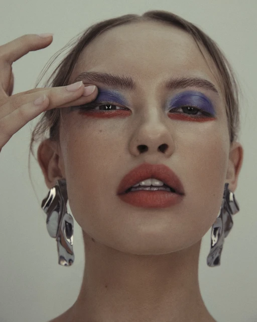 Close-up beauty shot of a woman with red lipstick and dramatic blue and red eye makeup, wearing large silver statement earrings.
