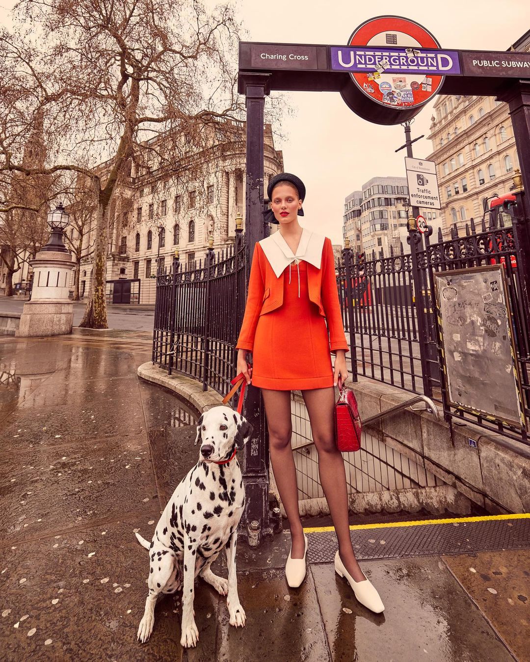 Stylish woman in an orange suit stands with a dalmatian dog near a London Underground entrance on a city street, showcasing fashion.