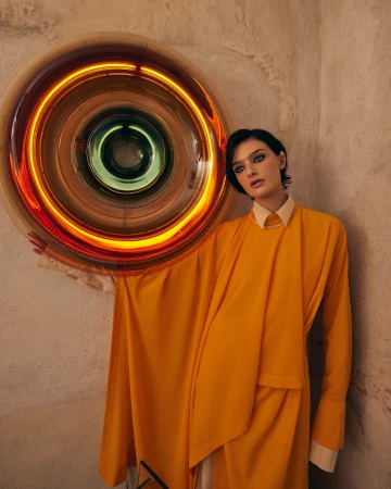 Stylish woman in yellow dress poses next to a round light art installation on a textured wall in an indoor setting. Fashion portrait.