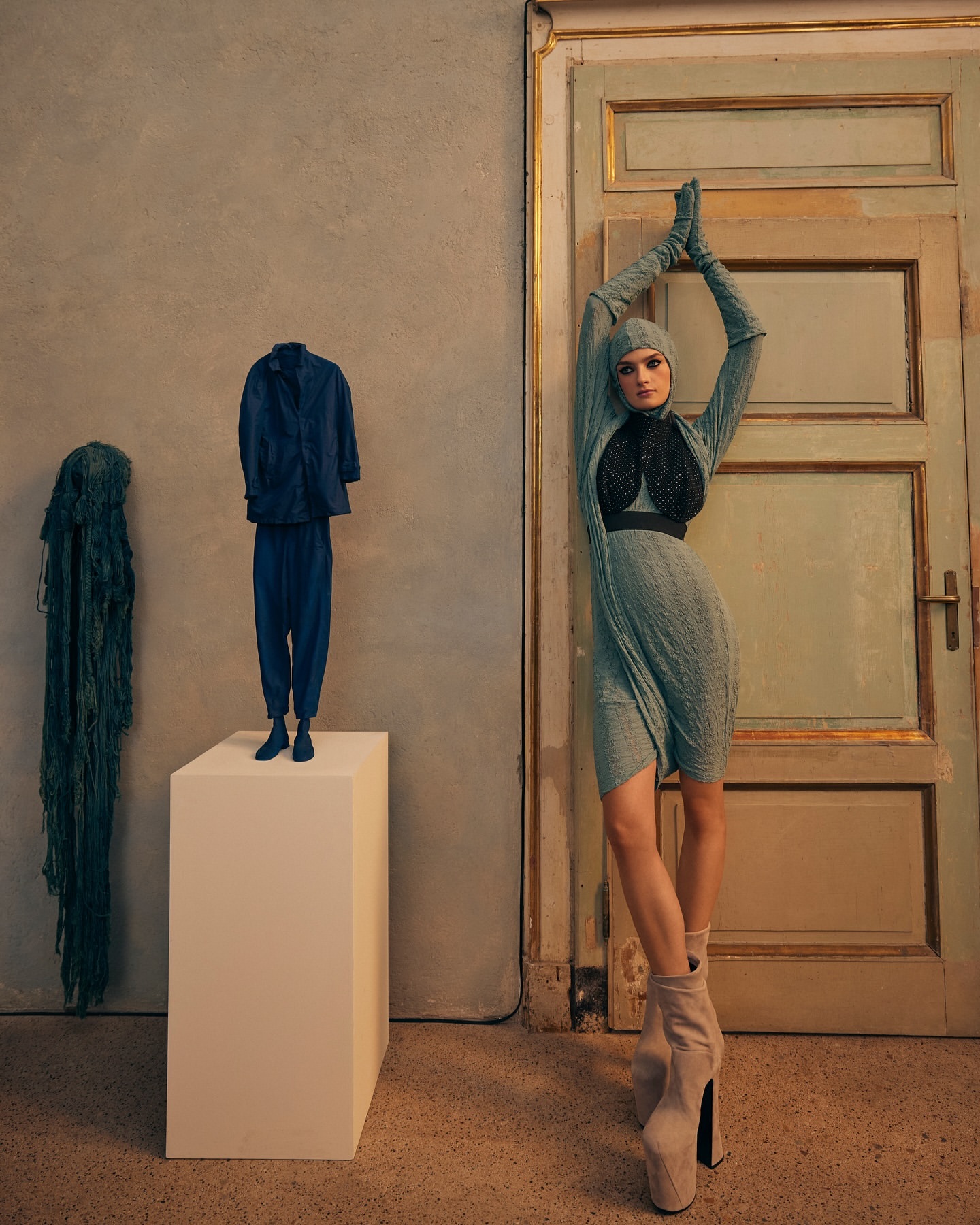 A woman poses in a blue dress, gloves and head covering near an old door with a blue suit and draped fabric on a white plinth.