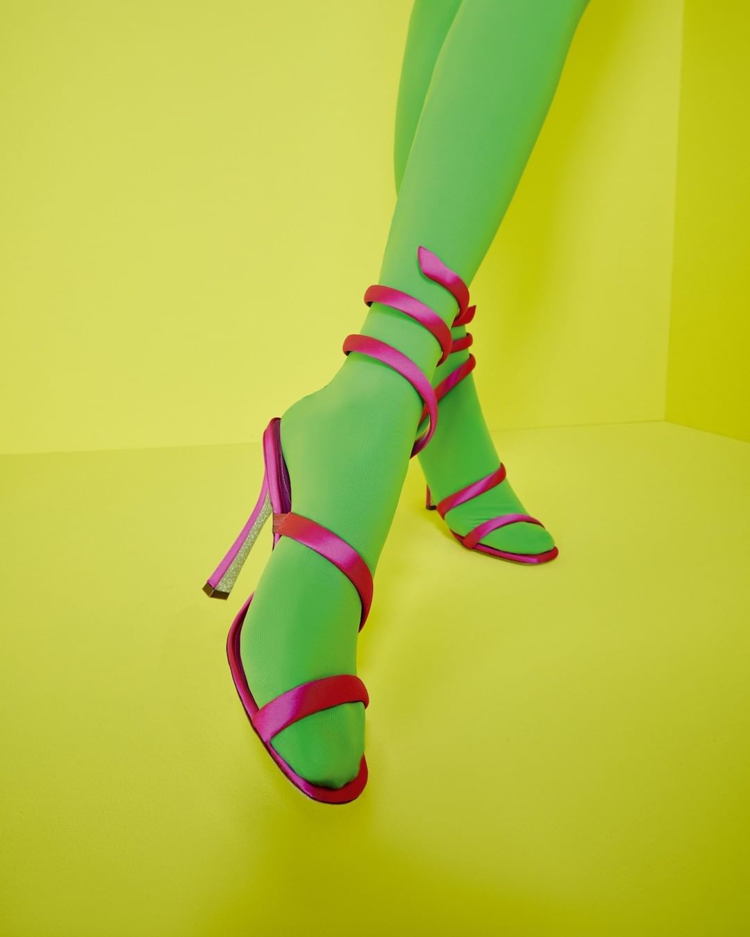 Close-up of legs wearing green tights and pink strappy high heel sandals on a yellow background creating a bright, vibrant fashion shot.