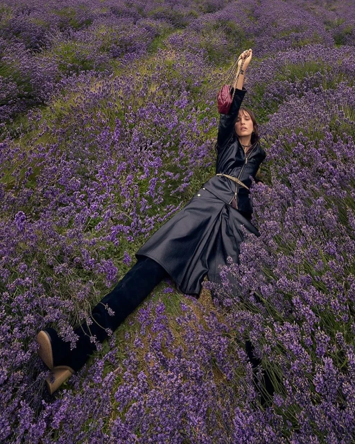Adult female model in black dress and boots lies in a lavender field with a purse, posing with one arm raised and looking at the camera.