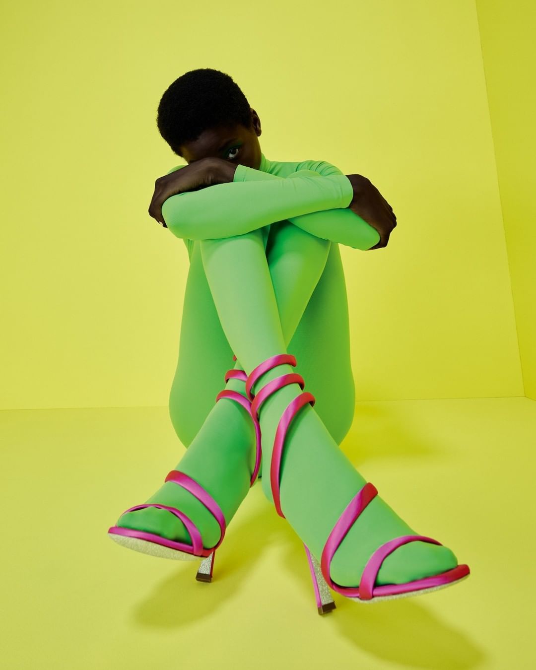 Woman in a lime green bodysuit and strappy pink heels sitting on a yellow backdrop in a studio for a fashion editorial shot.