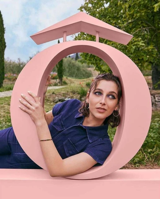 Eye-level shot of an adult female in a blue jumpsuit leaning against a pink circular sculpture in a lush outdoor setting.