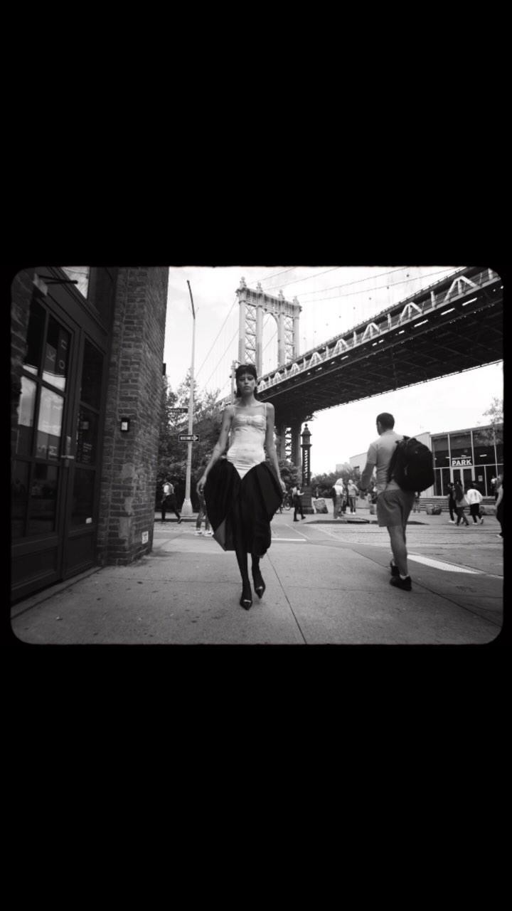 Black and white shot of a woman in a corset and large skirt walking on a city street, with a bridge visible in the background.