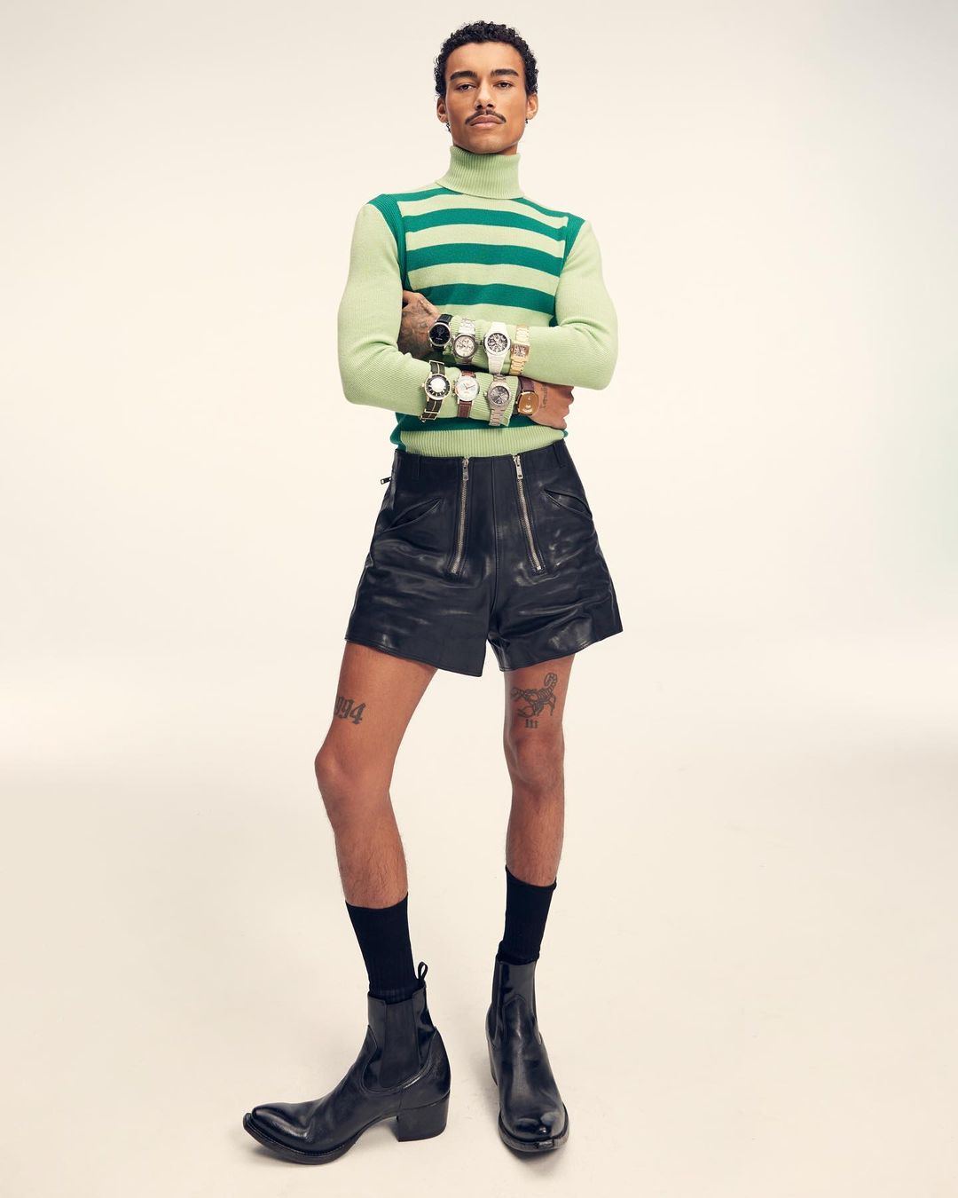 Man in striped turtleneck and leather shorts poses with multiple wristwatches, showcasing fashion and personal style against a neutral backdrop.