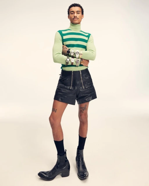 Man in striped turtleneck and leather shorts poses with multiple wristwatches, showcasing fashion and personal style against a neutral backdrop.