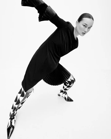 Model in black dress and patterned boots posing dramatically against a white seamless background