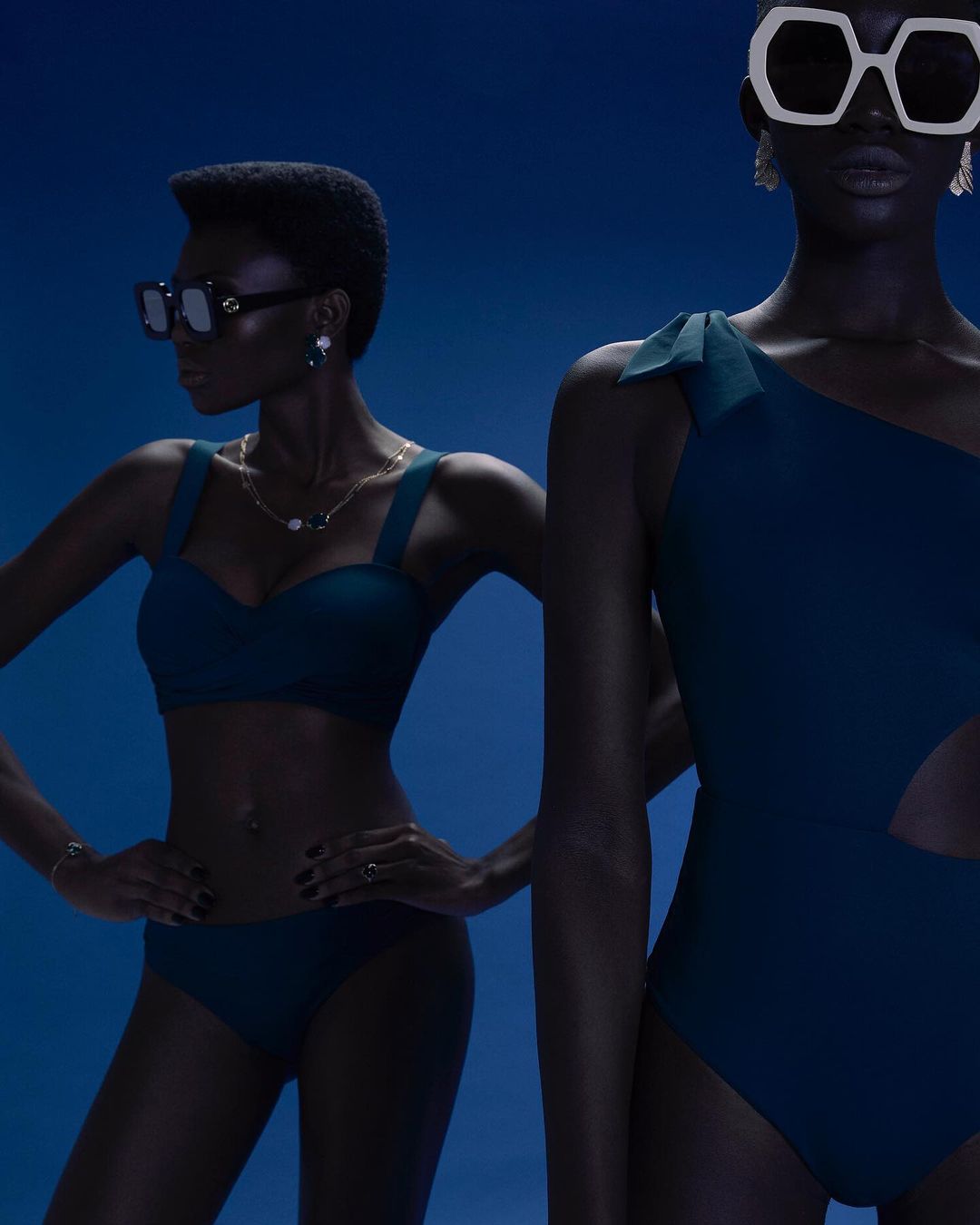 Two dark-skinned women pose in blue swimwear against a blue backdrop, one in a bikini and sunglasses, the other in a one-piece with statement glasses.