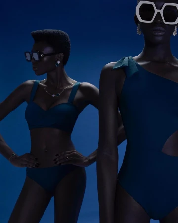 Two dark-skinned women pose in blue swimwear against a blue backdrop, one in a bikini and sunglasses, the other in a one-piece with statement glasses.