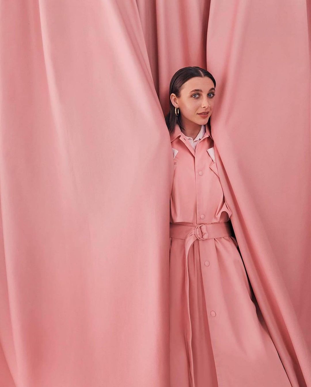 A woman with brown hair poses wearing a pink dress, framed by pink curtains, standing indoors with a gentle, natural-light illumination.