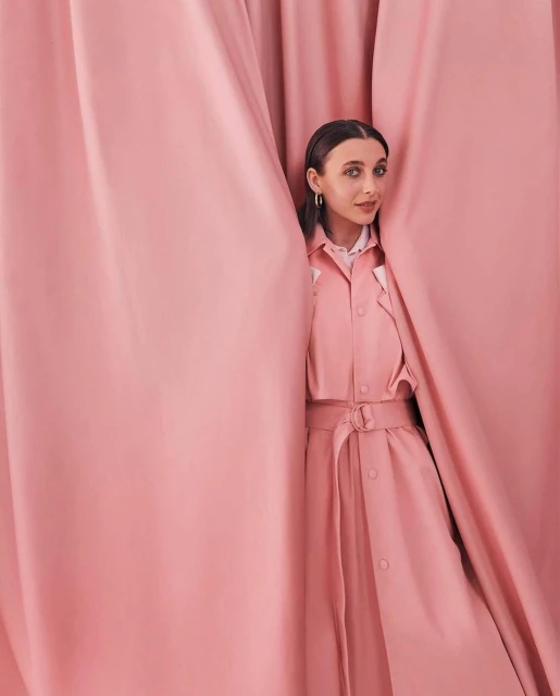 A woman with brown hair poses wearing a pink dress, framed by pink curtains, standing indoors with a gentle, natural-light illumination.