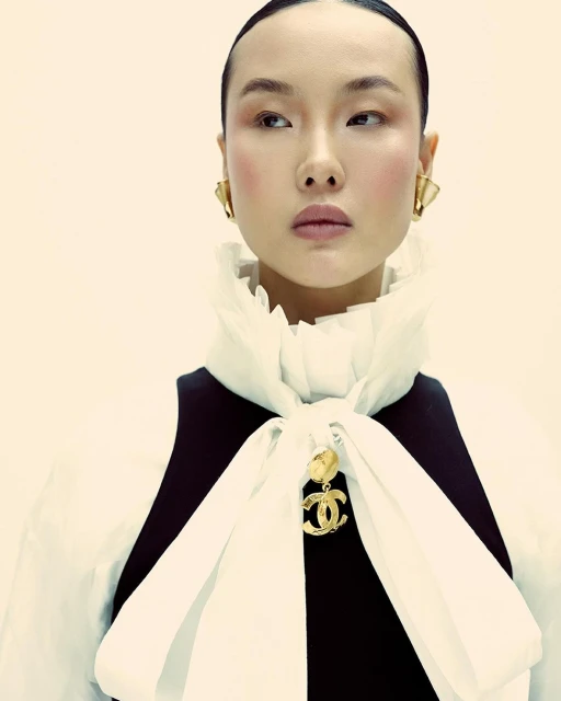 Headshot of a woman with pale skin, wearing gold earrings and a black and white ruffled collar dress, against a pale background.