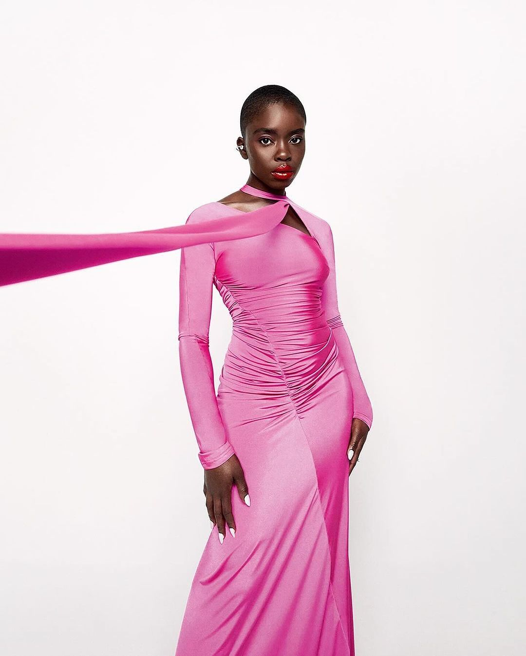 Elegant African woman in a long-sleeve hot pink gown poses in a studio, against a white backdrop, showing off a fashionable design.