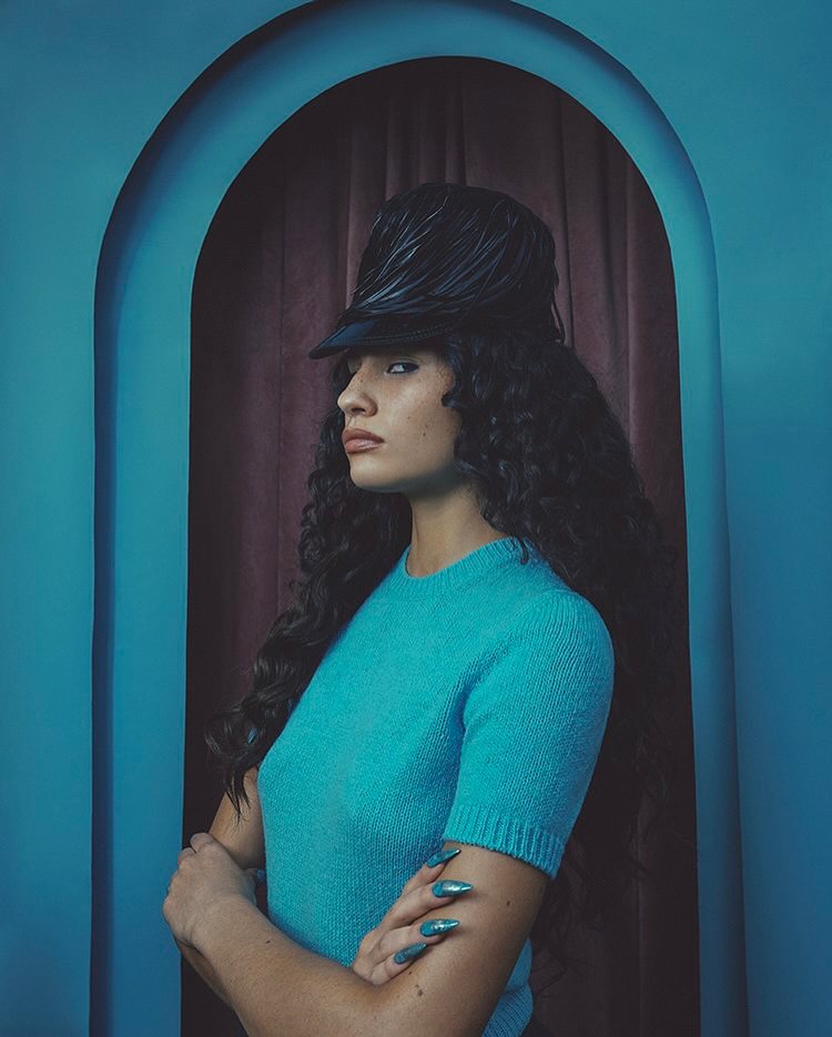 Portrait of a woman in a teal sweater and feathered hat, posing in front of an arched doorway with a maroon curtain backdrop.
