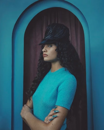 Portrait of a woman in a teal sweater and feathered hat, posing in front of an arched doorway with a maroon curtain backdrop.
