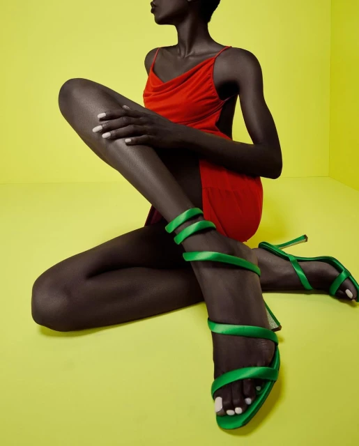 Fashion shot of an African woman in a red dress, sitting down with green heels, against a yellow background in studio lighting.