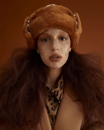 Model wearing a brown fluffy faux-fur ushanka hat, leopard-print shirt, and freckled makeup against a solid warm brown background