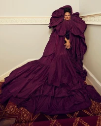 Woman in an oversized dramatic plum ruffled gown with a sweeping train standing indoors photographed by Carla Guler