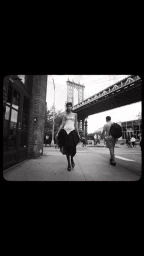 Black and white street photography by Carla Guler showing a model walking on a city sidewalk beneath a large suspension bridge.
