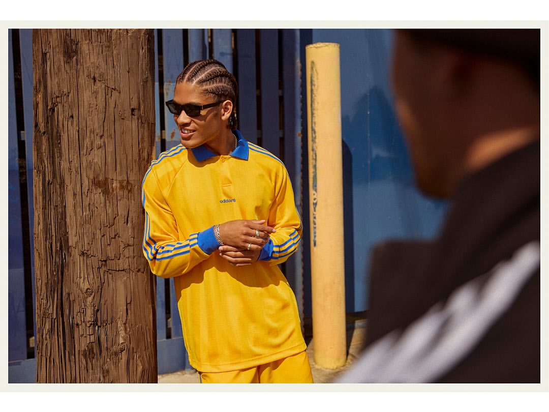 A young adult male with cornrow braids wears sunglasses and a yellow Adidas collared shirt, standing in front of a blue wall outdoors.