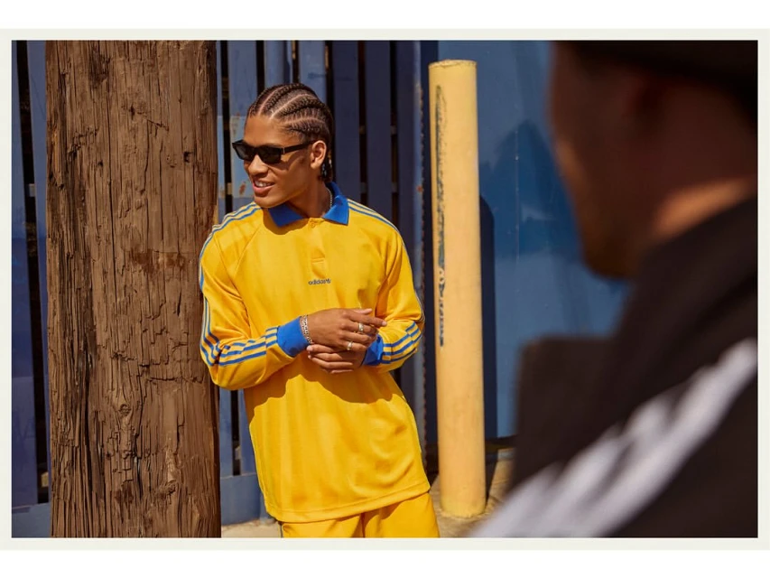 A young adult male with cornrow braids wears sunglasses and a yellow Adidas collared shirt, standing in front of a blue wall outdoors.