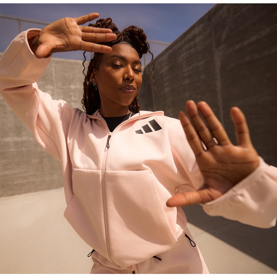 Athletic woman in a pink Adidas tracksuit poses with hands framing her face against a neutral concrete wall in an outdoor setting.