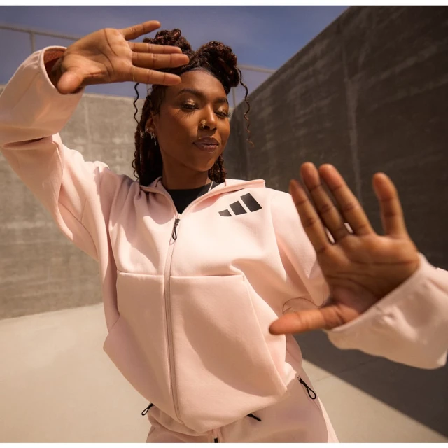Athletic woman in a pink Adidas tracksuit poses with hands framing her face against a neutral concrete wall in an outdoor setting.